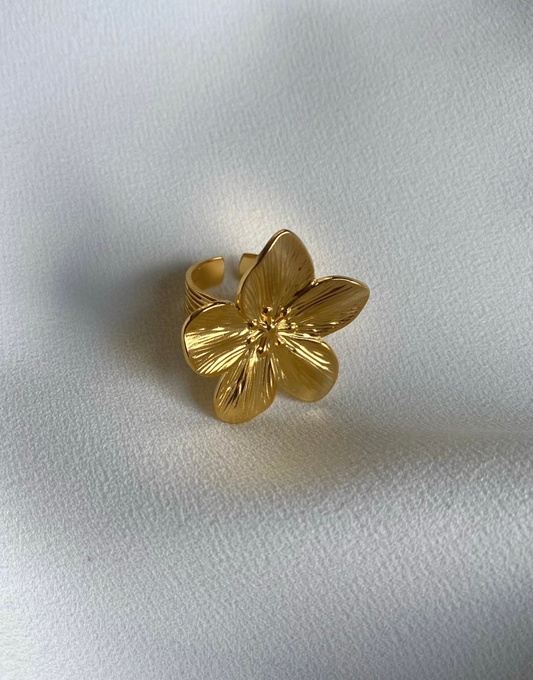 A stunning gold ring featuring a large, intricately designed flower with detailed petals. The ring is adjustable, showcasing its chunky and bold design, perfect for making a statement. The shiny finish highlights its luxurious appeal, set against a soft, elegant fabric background.
