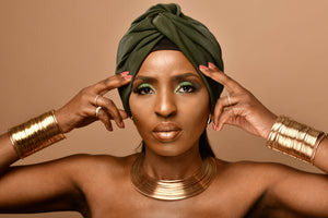 Portrait of a woman with dramatic metallic jewellery, featuring a wide silver neckpiece and cylindrical arm cuffs. Her elegant makeup highlights her confident expression against a peach background.