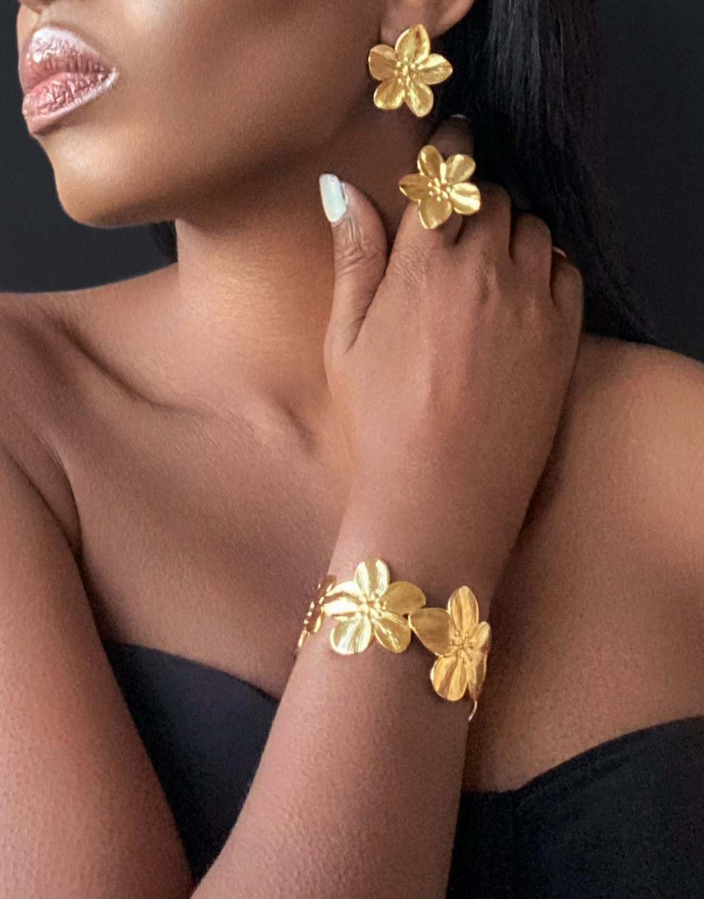 Elegant close-up showcasing a model wearing matching gold flower-shaped earrings, ring and a floral bangle bracelet. The jewellery's intricate design and polished finish exude sophistication, perfect for elevating any outfit. The black backdrop highlights the luxurious gold tones, emphasizing the timeless beauty of these statement accessories.