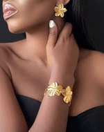 Cargar imagen en el visor de la galería, Elegant close-up showcasing a model wearing matching gold flower-shaped earrings and a floral bangle bracelet. The jewellery&#39;s intricate design and polished finish exude sophistication, perfect for elevating any outfit. The black backdrop highlights the luxurious gold tones, emphasizing the timeless beauty of these statement accessories.
