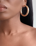 Cargar imagen en el visor de la galería, Close-up of a woman wearing elegant Cairo Large Clip-On Hoop Earrings in premium gold-plated brass, showcasing their substantial size and glossy finish. The earrings add a touch of sophistication to her poised appearance.
