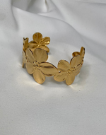 Cargar imagen en el visor de la galería, Side view of the ABYDOS Flower Cuff Bracelet, showcasing its intricate floral design in 18k gold plating over stainless steel, beautifully positioned against a soft white fabric background.
